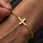 A close-up of a gold chain bracelet with a cross pendant engraved, worn on a person's wrist with fingers adjusting it.