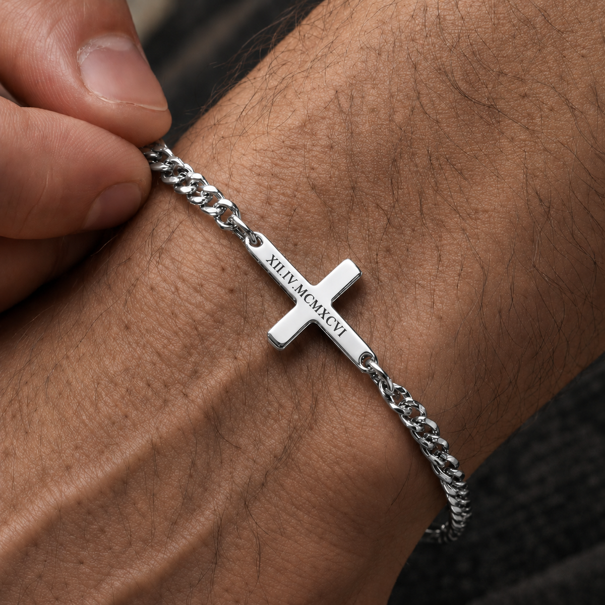A silver-toned chain bracelet with a cross charm engraved, worn on a person's wrist, with fingers adjusting the chain.