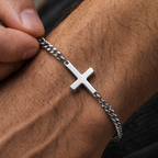A silver cross pendant bracelet on a silver chain, shown worn on a person's wrist with fingers adjusting it.