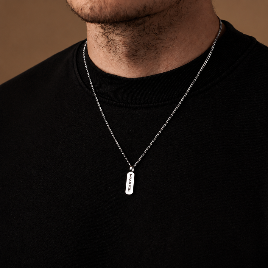 A close-up of a man wearing a silver chain necklace with a rectangular silver pendant engraved with the word 'MORAL'.