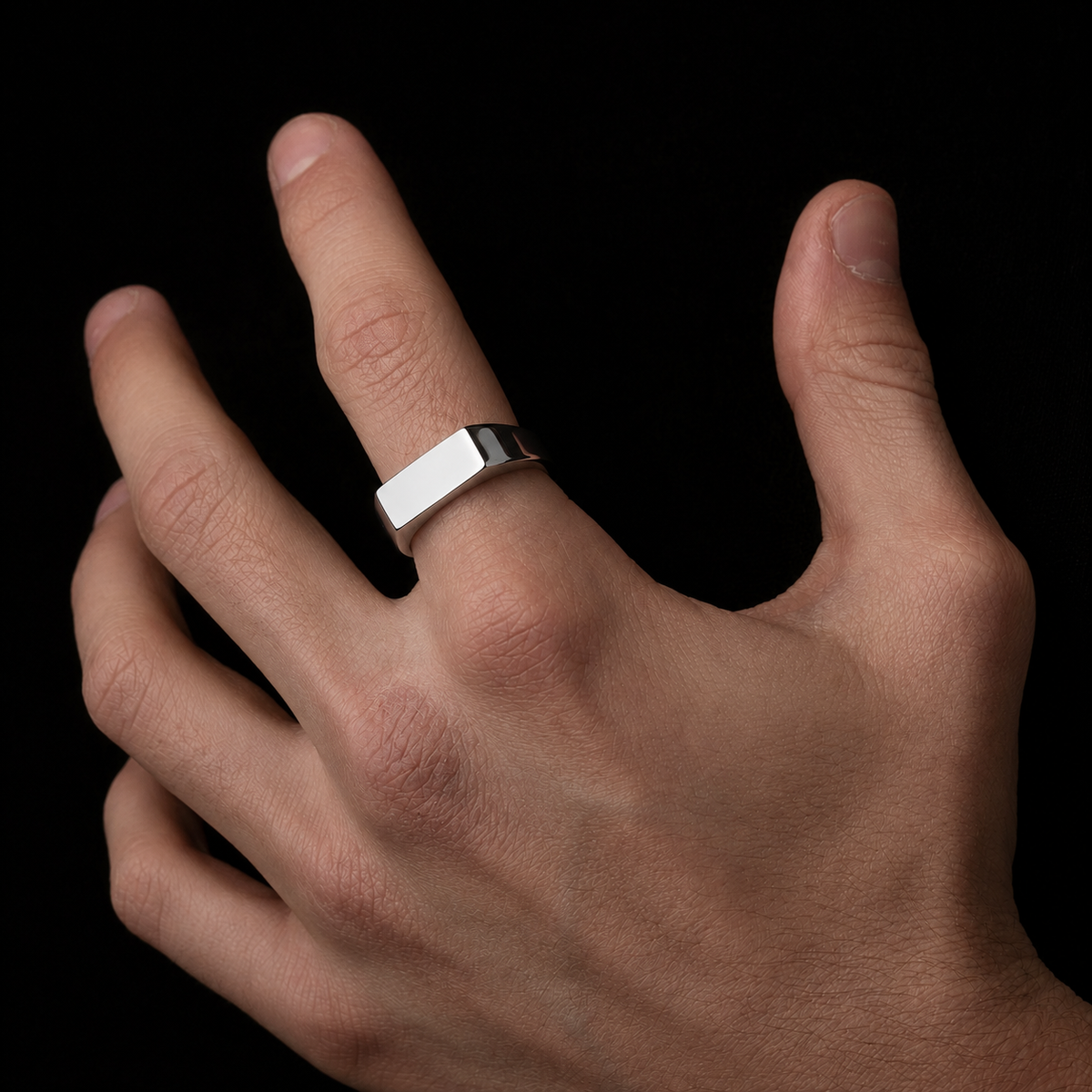 A silver-toned metal ring with a flat, rectangular bezel setting worn on a person's ring finger, shown against a solid black background.