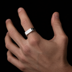 A silver-toned metal ring with a flat, rectangular bezel setting worn on a person's ring finger, shown against a solid black background.