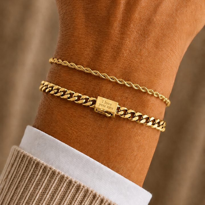 Two gold bracelets, one a delicate cable chain and the other a chunkier curb link with a small engraved square charm, worn on a person's wrist over a beige knit sweater cuff.