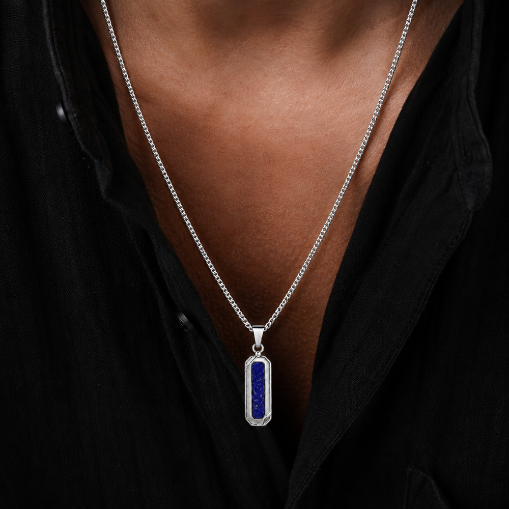 A close-up of a person wearing a silver chain necklace with a vertical rectangular pendant featuring a deep blue lapis lazuli stone in a silver bezel setting, worn over a black shirt.