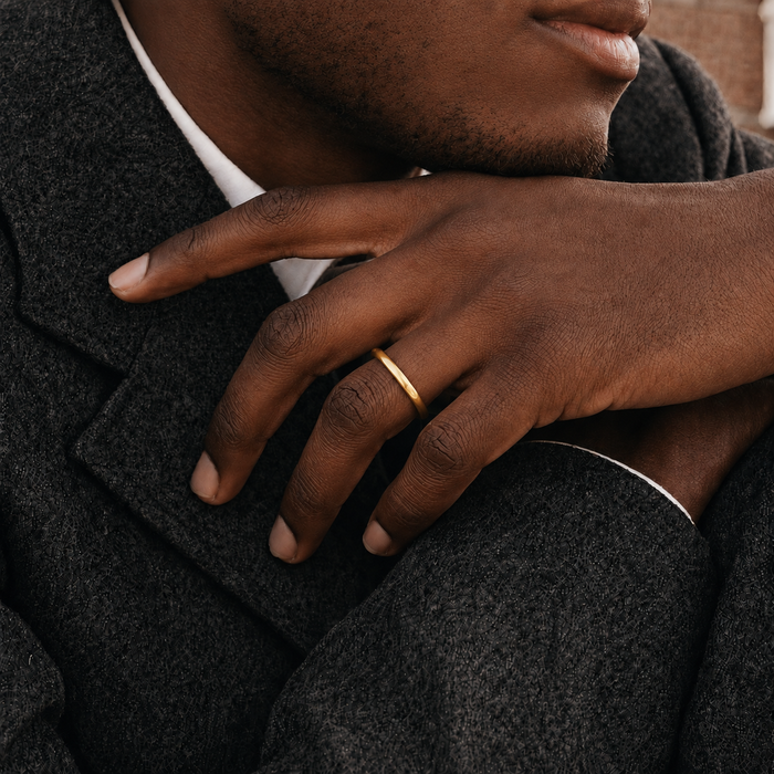 A close-up lifestyle shot of a man wearing a simple, slim gold band ring on his ring finger, dressed in a dark gray wool coat over a white collared shirt.