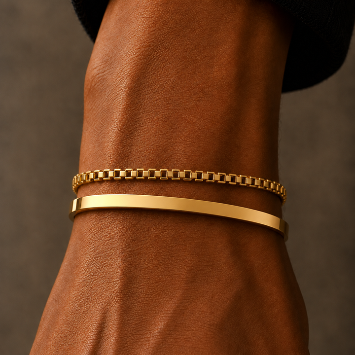 Two gold bracelets worn on a person's wrist, featuring a thin box-link chain bracelet and a thicker polished bar bracelet, shown against a warm brown skin tone and dark background.