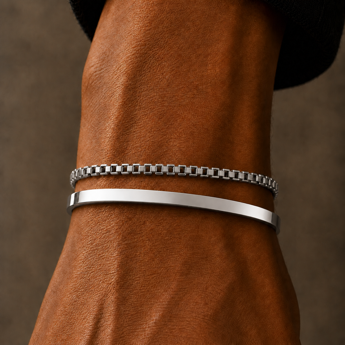 Two silver-toned metal bracelets worn on a person's wrist, with one being a thin chain bracelet and the other a smooth bangle, shown against a warm brown leather background.
