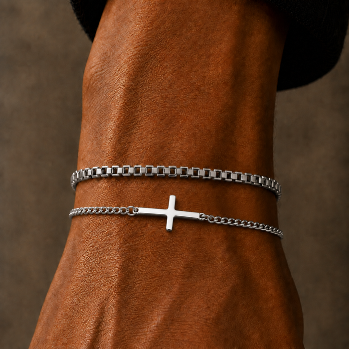 Two silver bracelets worn on a wrist: a thicker rectangular chain bracelet and a delicate chain bracelet with a small cross pendant, shown against a brown leather background.