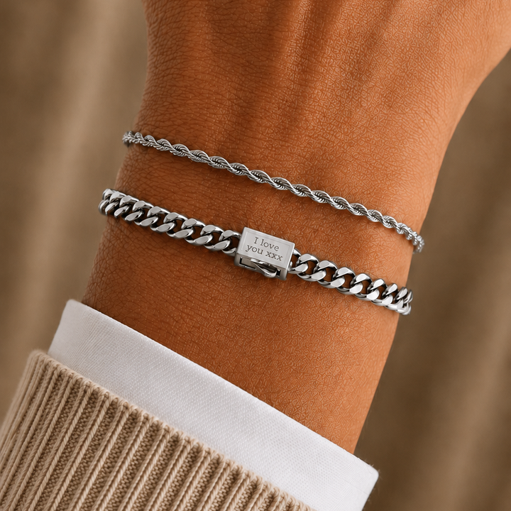 Two silver bracelets worn on a person's wrist, including a twisted chain bracelet and a curb link bracelet with a small engraved square charm, shown against a warm brown background.