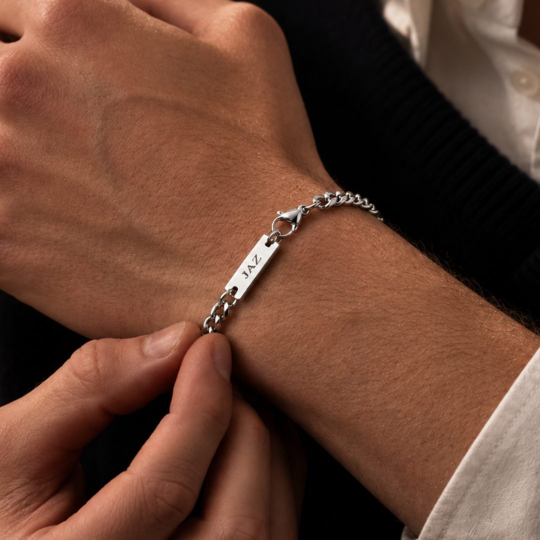 A close-up of a silver chain bracelet with a rectangular engraved plate worn on a person's wrist, shown against a dark sweater and white collared shirt.