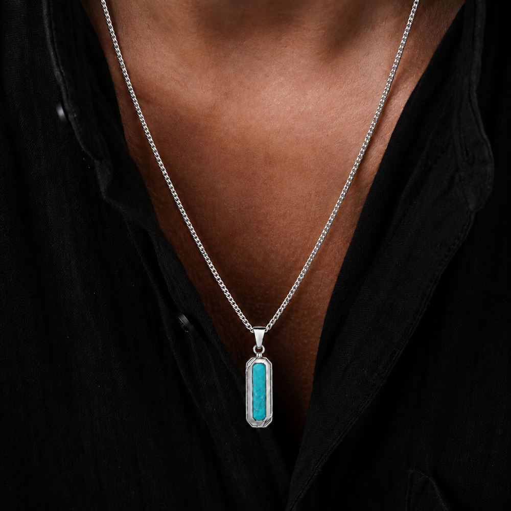 A close-up of a person wearing a silver chain necklace with a vertical rectangular pendant featuring a bright turquoise center set in a silver frame, worn over a black shirt.