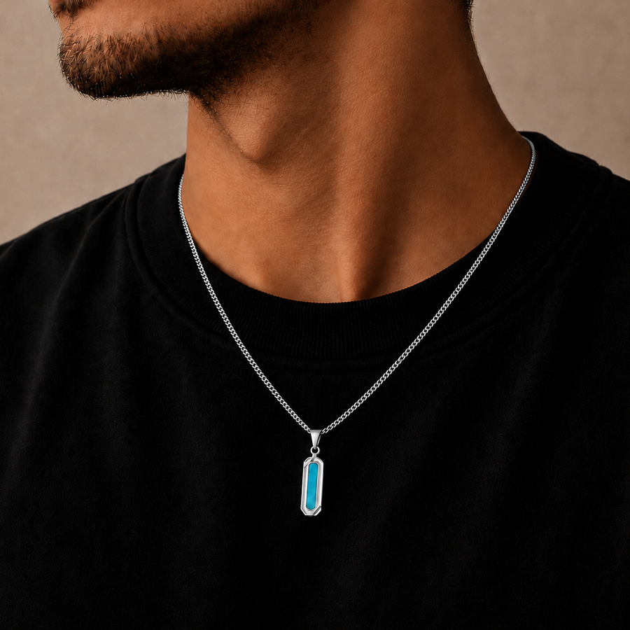 A close-up of a man wearing a silver chain necklace with a vertical rectangular pendant featuring a bright turquoise blue center, set against a plain beige background.