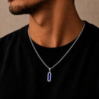 A close-up of a man wearing a silver chain necklace with a vertical rectangular pendant featuring a deep blue center and silver borders, set against a plain beige background.