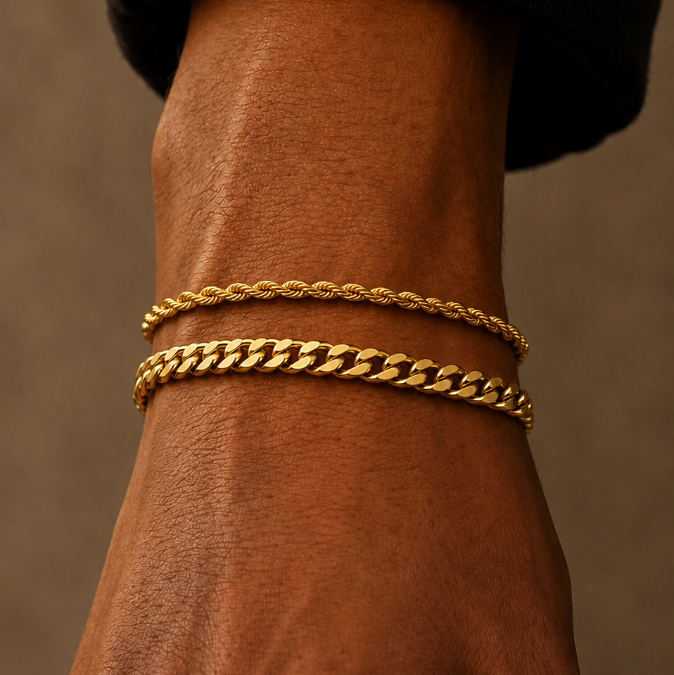 Two gold bracelets, one a thin twisted rope style and the other a chunky curb link style, worn stacked on a person's wrist against a warm brown background.
