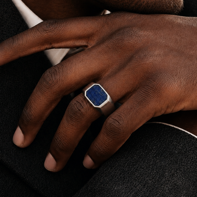 A close-up of a man's hand resting on his lap, wearing a silver-toned signet ring with a square blue stone.