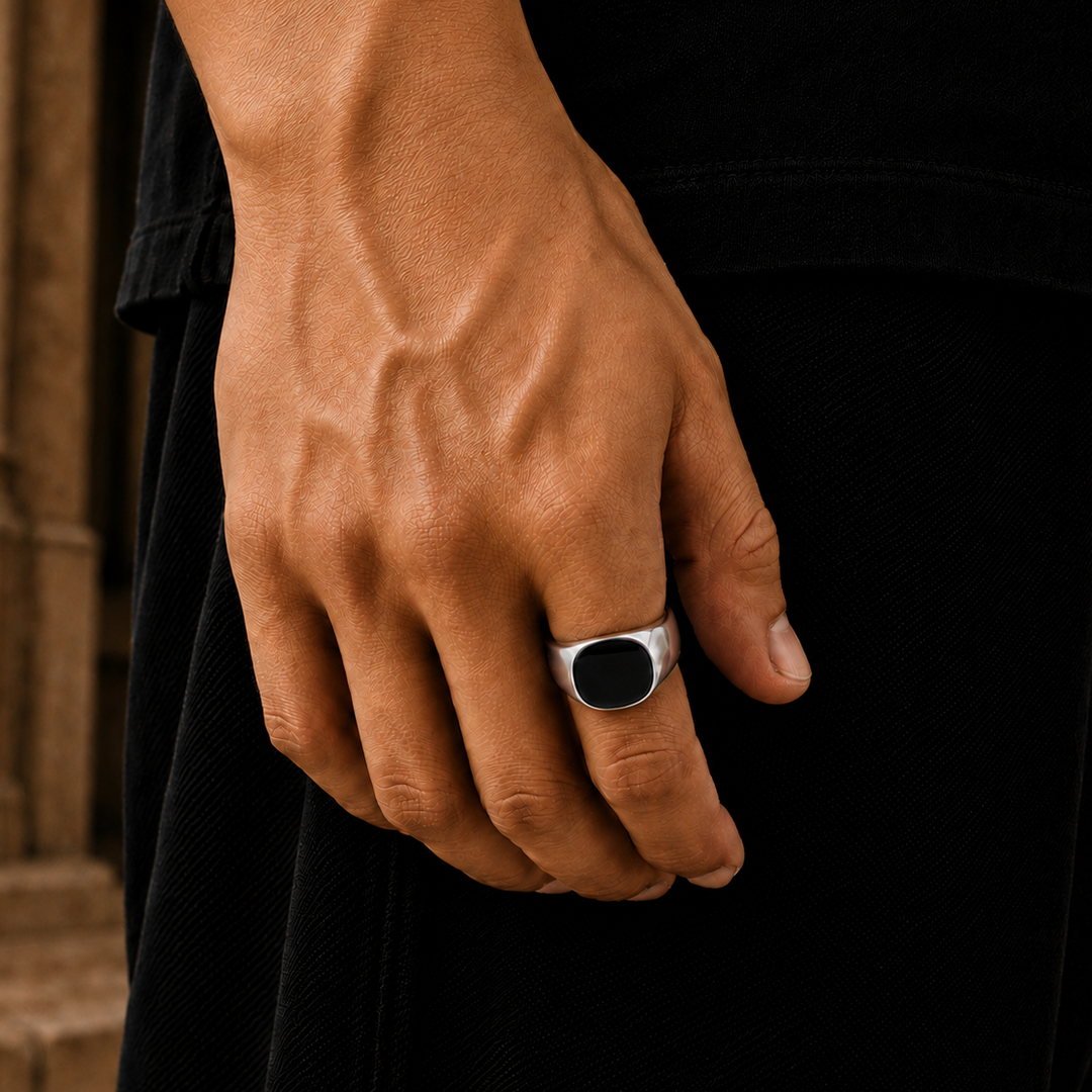 A close-up of a man's hand wearing a silver signet ring with a black oval face, set against a dark shirt and stone architectural background.