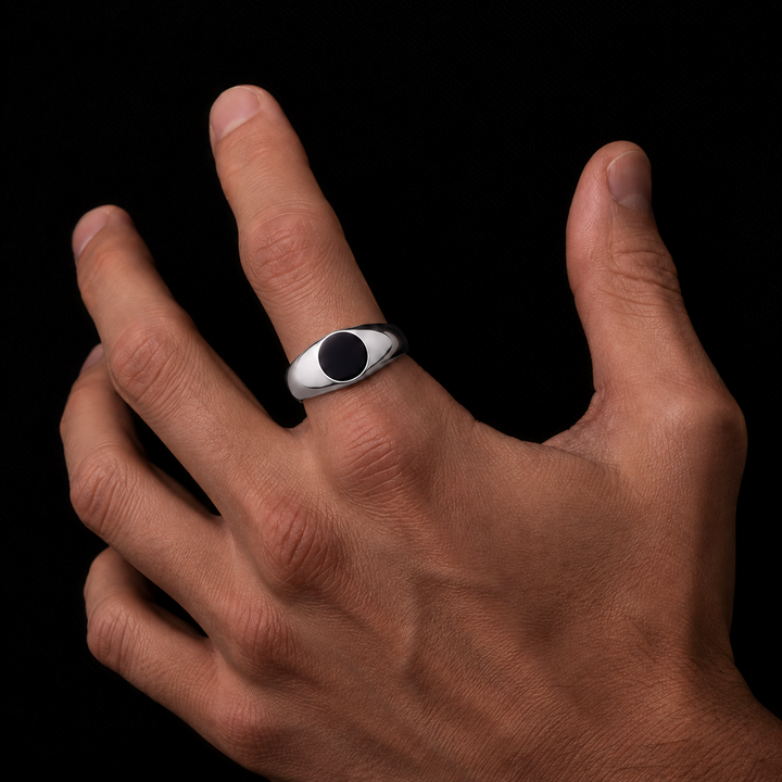 A silver-toned men's signet ring with a black oval inlay, shown worn on a person's ring finger against a solid black background.