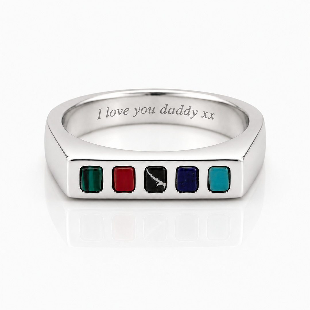 A silver-toned metal ring shaped like a tablet, featuring five small colored square inlays along the front edge: green, red, black with a white sword engraving, dark blue, and teal. The inside of the band is engraved with the text 'I love you daddy xx'.