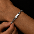 A close-up of a silver bracelet being adjusted on a person's wrist. The bracelet features a braided chain section and a flat rectangular nameplate engraved with the name 'STANLEY'.
