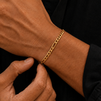 A close-up of a man's wrist wearing a gold chain link bracelet, with his other hand resting on his chest. He is wearing a black button-down shirt.