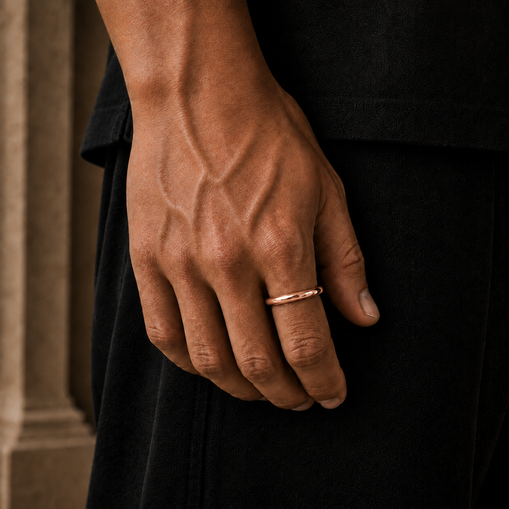 A close-up of a person's hand wearing a thin rose gold band ring, set against a dark black garment and a blurred beige background.