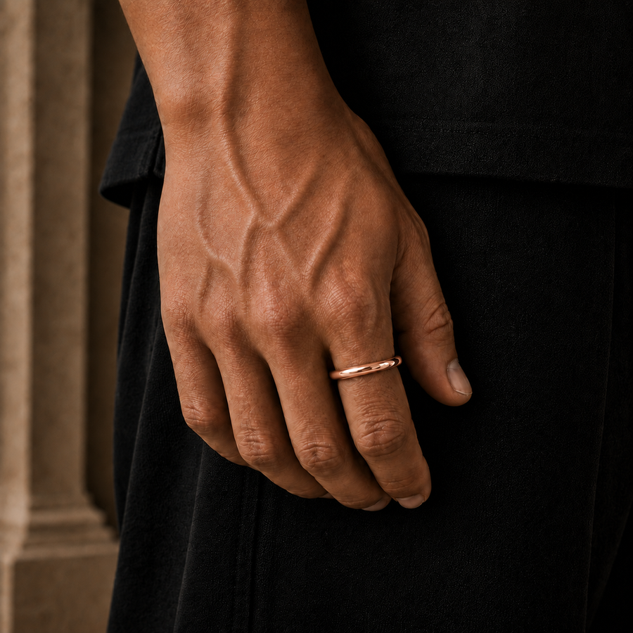A close-up of a person's hand wearing a thin rose gold band ring, set against a dark black garment and a blurred beige background.