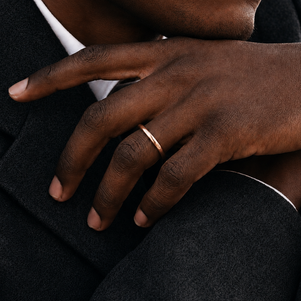 A minimalist rose gold band ring worn on a man's ring finger, shown in a close-up lifestyle shot against a dark suit jacket and white shirt.
