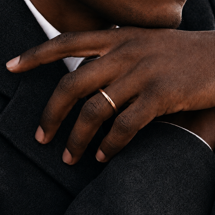 A minimalist rose gold band ring worn on a man's ring finger, shown in a close-up lifestyle shot against a dark suit jacket and white shirt.