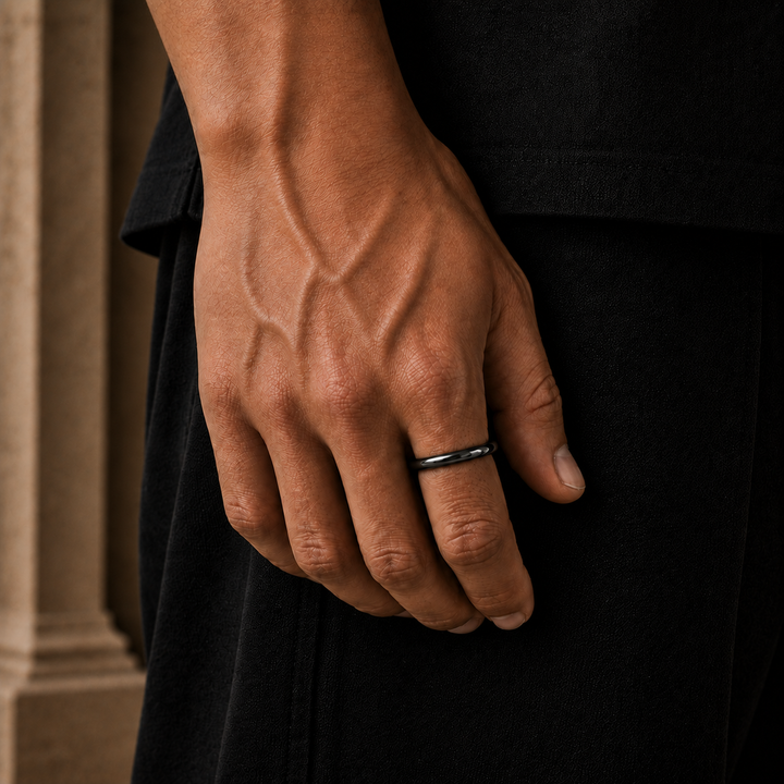 A close-up of a man's hand wearing a simple black metal band ring, dressed in a black shirt and dark trousers.