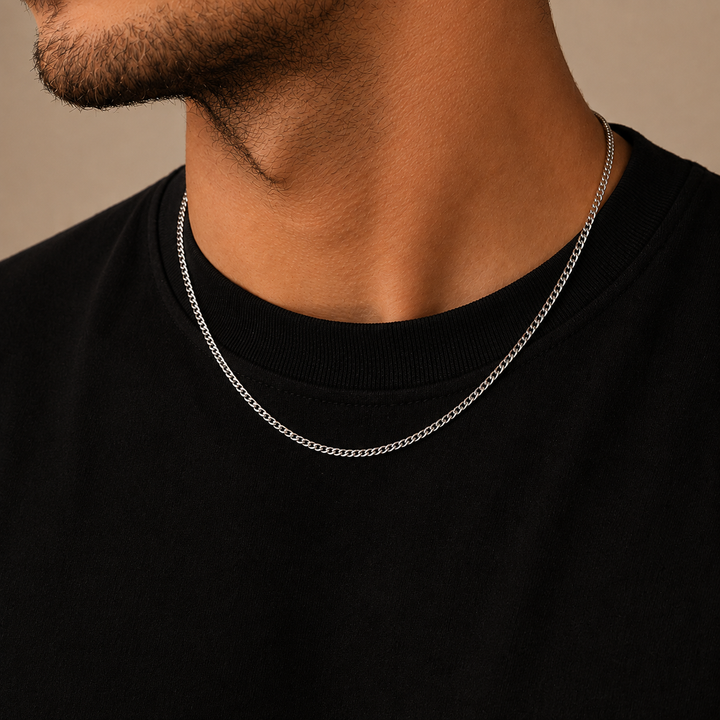 A close-up of a man's neck and upper chest wearing a thin silver chain necklace over a black crewneck shirt, against a warm beige background.