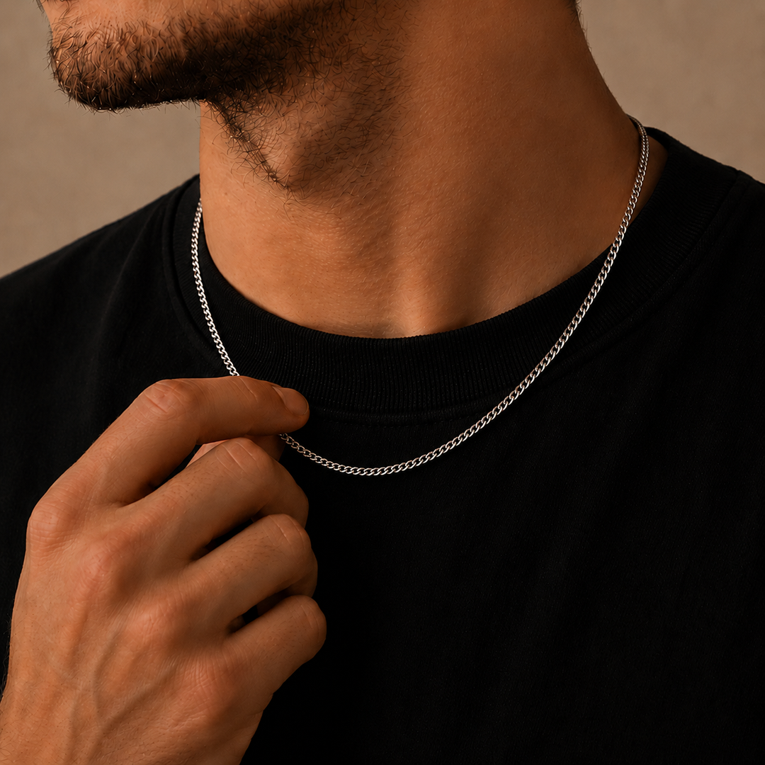 A close-up of a man wearing a thin silver chain necklace over a black crewneck shirt, with his hand resting near the collar.