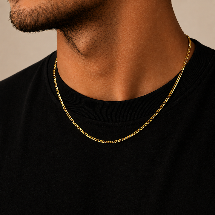 A close-up of a man's neck and lower face wearing a delicate gold chain necklace over a black crewneck shirt, set against a warm beige background.