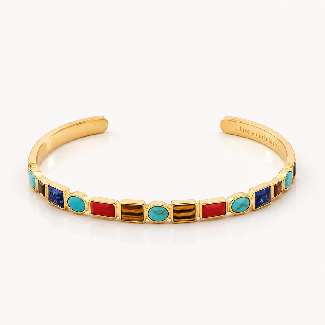 A gold cuff bracelet featuring a row of colorful rectangular and oval bezel-set stones in red, turquoise, blue, and brown, displayed on a clean white background.