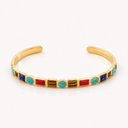 A gold cuff bracelet featuring a row of colorful rectangular and oval bezel-set stones in red, turquoise, blue, and brown, displayed on a clean white background.