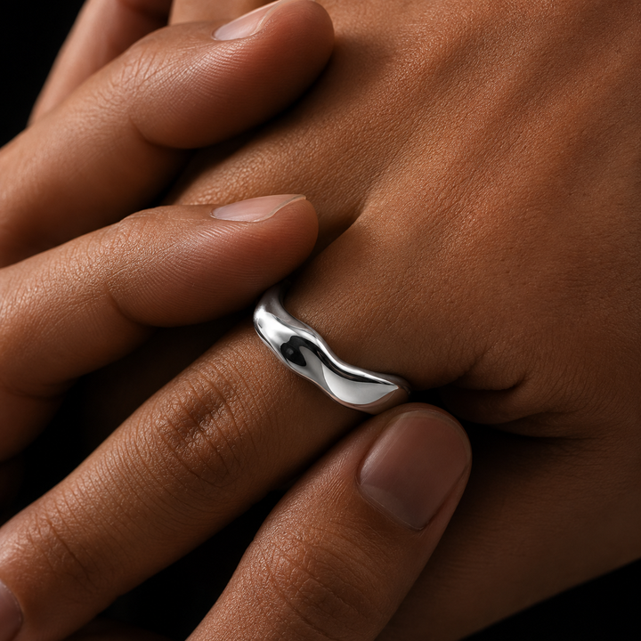 A close-up of a hand wearing a polished silver ring with a wavy, fluid design, set against a dark background.
