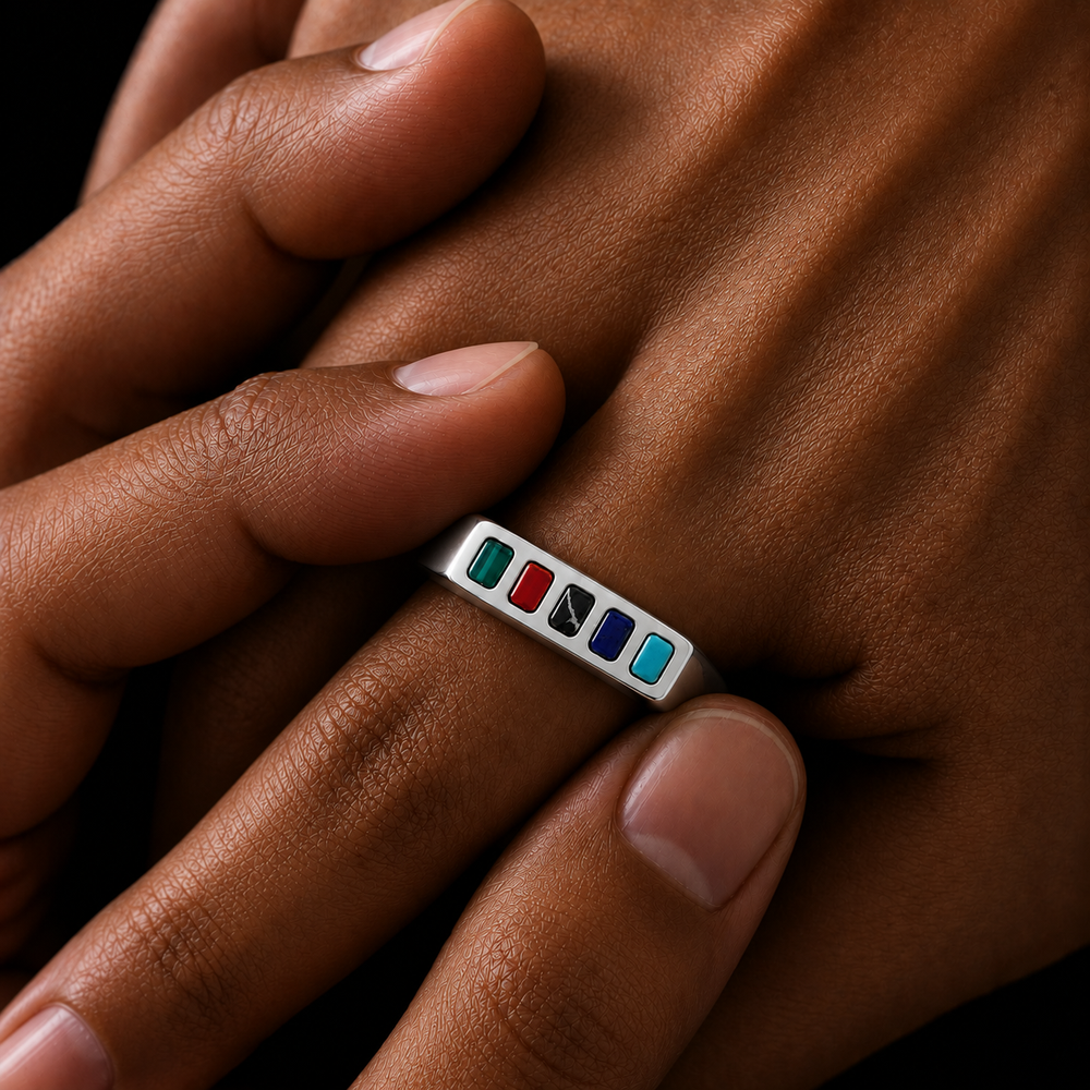A close-up of a silver-toned ring with six small rectangular inlay stones in green, red, black, white, blue, and light blue, worn on a person's finger against a dark background.