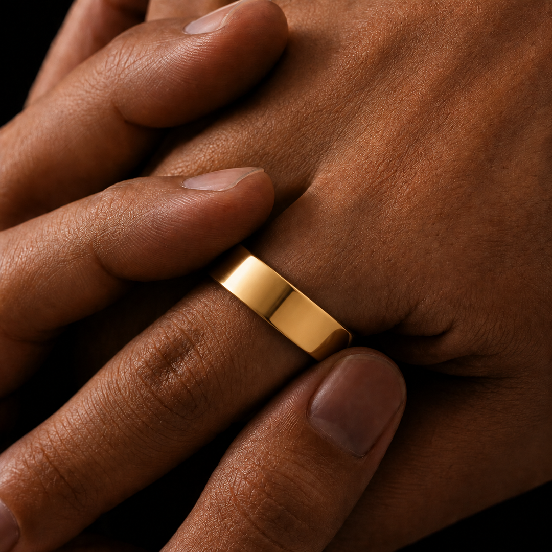 A close-up of a polished yellow gold band ring worn on a finger, shown against a person's skin with a dark background.