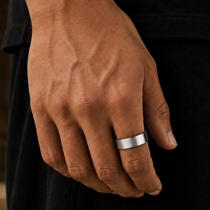 A close-up of a person's hand wearing a simple, polished silver band ring, set against a dark shirt and a blurred stone background.