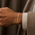 A close-up of a person's wrist wearing a thin, textured gold chain bracelet, paired with a brown textured jacket and a white dress shirt cuff.
