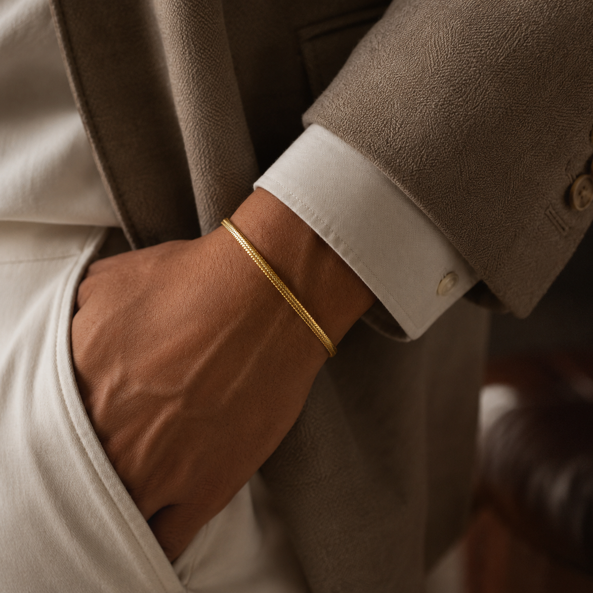 A close-up of a person's hand and wrist wearing a thin, polished gold snake chain bracelet, styled with a white dress shirt cuff and a textured taupe blazer.