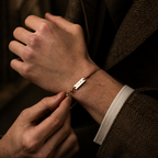 A close-up lifestyle shot of a man's hands adjusting a delicate gold chain bracelet with a rectangular engraved plate that reads 'LOVE' on his wrist. He is wearing a brown herringbone tweed jacket and a white dress shirt cuff.