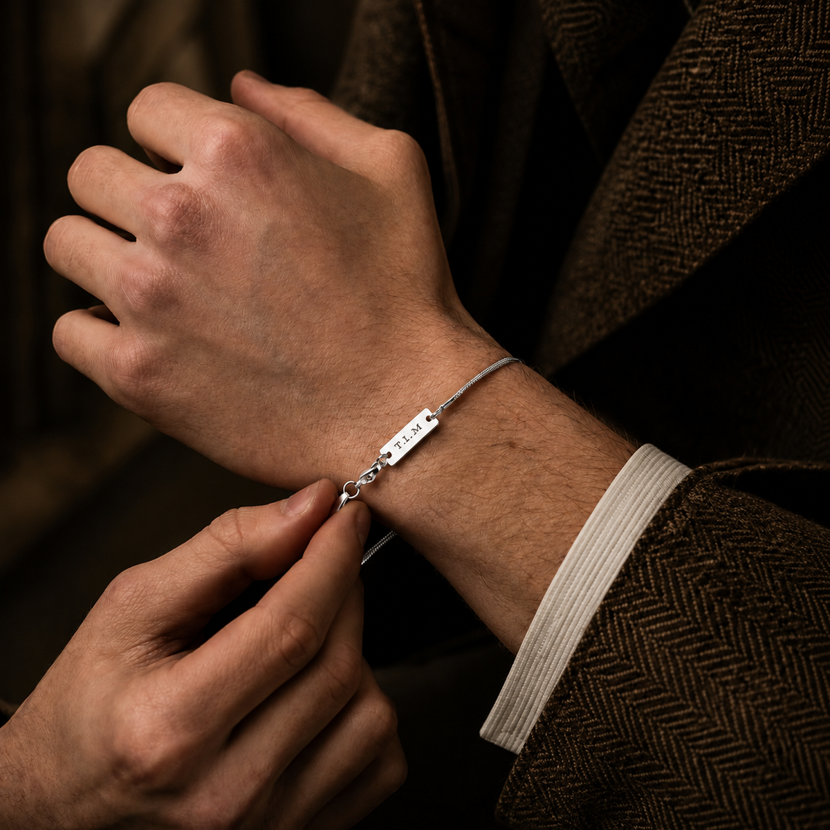 A close-up of a person's wrist wearing a slim silver bracelet with a rectangular engraved plate that reads 'I LOVE YOU'. The person is wearing a brown herringbone jacket and a white dress shirt cuff.