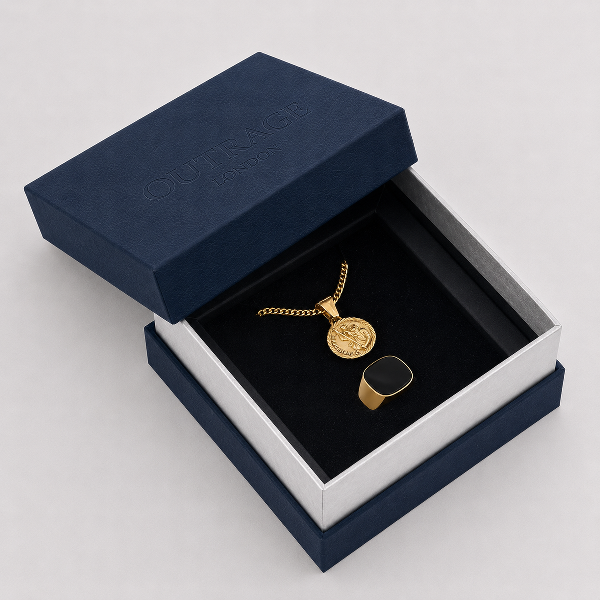 A gold coin pendant necklace and matching ring displayed inside an open navy blue jewelry box with a black velvet insert. The embossed brand name 'OUTRAGE London' is visible on the navy lid.