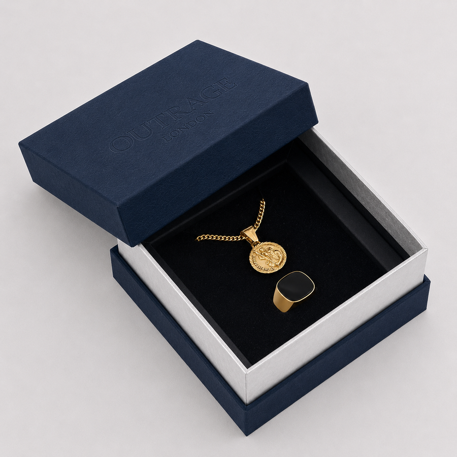A gold coin pendant necklace and matching ring displayed inside an open navy blue jewelry box with a black velvet insert. The embossed brand name 'OUTRAGE London' is visible on the navy lid.