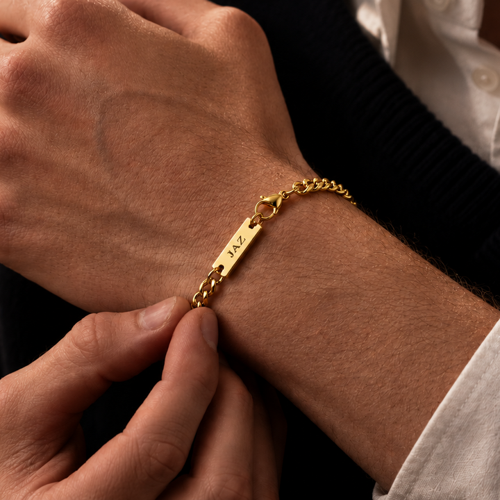 A close-up of a gold chain bracelet with a rectangular engraved plate worn on a person's wrist, shown with hands adjusting it over a dark sweater and white collared shirt.