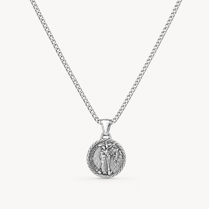 A silver chain necklace with a round, detailed pendant featuring a religious figure, shown against a light off-white background.