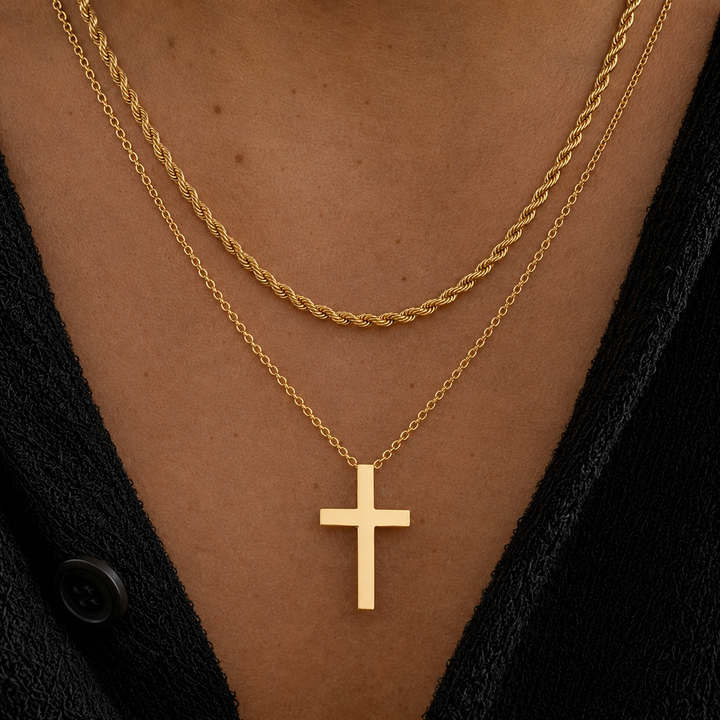 A close-up of a person's chest wearing two layered gold necklaces: a twisted rope chain and a delicate chain with a flat cross pendant, styled with a black knit top.