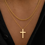 A close-up of a person's chest wearing two layered gold necklaces: a twisted rope chain and a delicate chain with a flat cross pendant, styled with a black knit top.