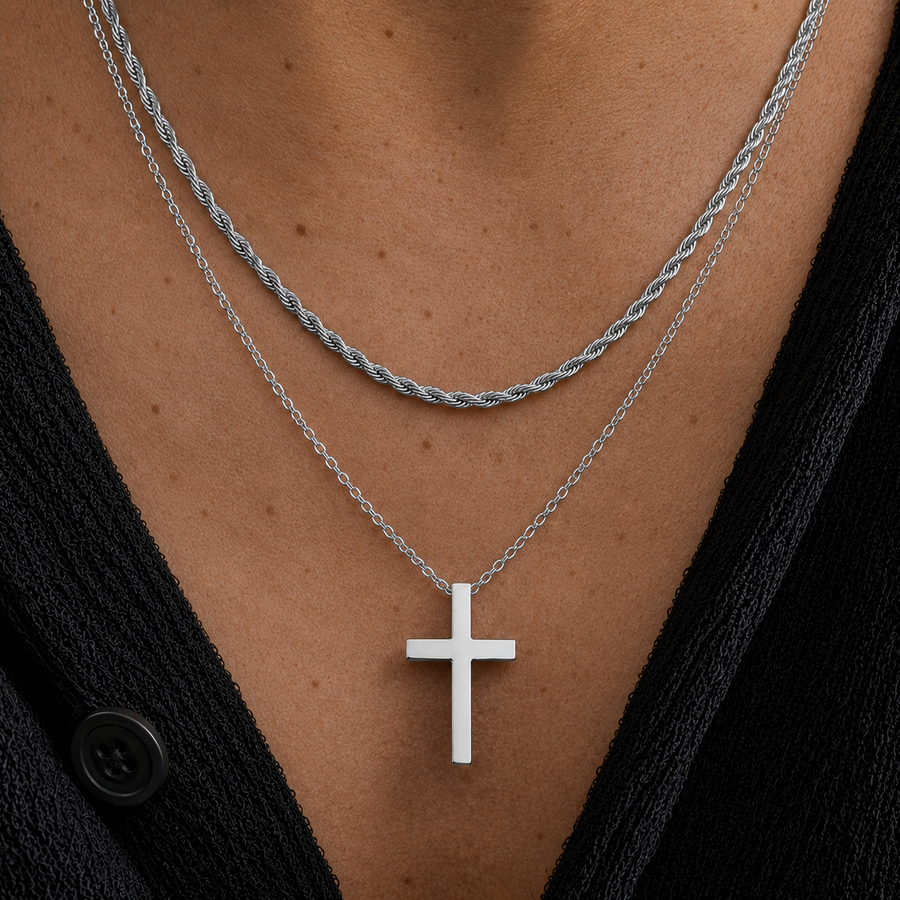 A close-up of a person's chest wearing two layered necklaces: a silver twisted rope chain and a silver chain with a flat cross pendant, over a black top.