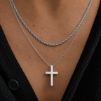 A close-up of a person's chest wearing two layered necklaces: a silver twisted rope chain and a silver chain with a flat cross pendant, over a black top.
